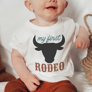 May include: A white baby onesie with a black bull silhouette and the text "my first rodeo" in teal and brown.
