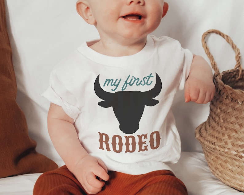 May include: A white baby onesie with a black bull silhouette and the text "my first rodeo" in teal and brown.