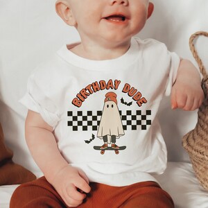 May include: White short-sleeved t-shirt with a graphic of a ghost wearing a red beanie and skateboarding. The text "Birthday Dude" is printed in orange above the ghost.