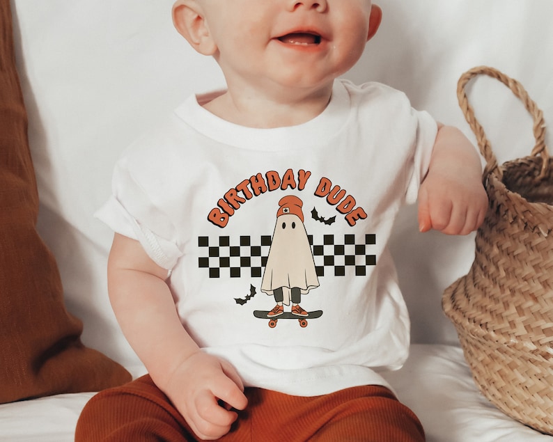May include: White short-sleeved t-shirt with a graphic of a ghost wearing a red beanie and skateboarding. The text "Birthday Dude" is printed in orange above the ghost.