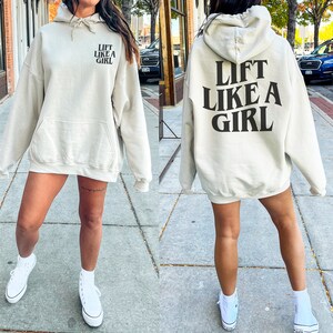 K&ouml;nnte beinhalten: Ein hellbeiger Hoodie mit einem schwarzen Aufdruck, der auf der R&uuml;ckseite "Lift Like A Girl" lautet und auf der Vorderseite denselben Text in kleinerer Schrift.