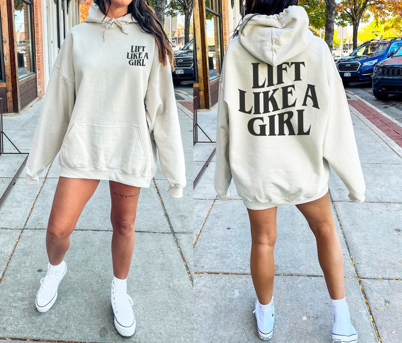 K&ouml;nnte beinhalten: Ein hellbeiger Hoodie mit einem schwarzen Aufdruck, der auf der R&uuml;ckseite "Lift Like A Girl" lautet und auf der Vorderseite denselben Text in kleinerer Schrift.