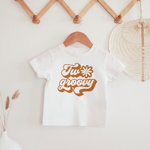 May include: White t-shirt with a brown graphic that says "Two groovy" with a daisy in the middle of the "o" in "two".