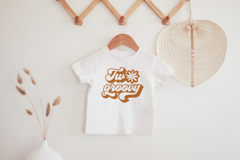 May include: White t-shirt with a brown graphic that says "Two groovy" with a daisy in the middle of the "o" in "two".