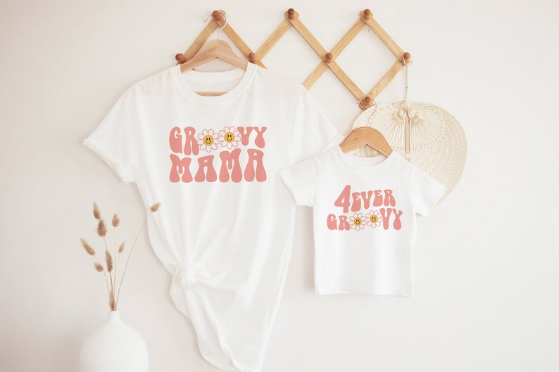 K&ouml;nnte beinhalten: Zwei wei&szlig;e T-Shirts, eine Erwachsenen- und eine Kindergr&ouml;&szlig;e, h&auml;ngen an Holzb&uuml;geln. Das T-Shirt f&uuml;r Erwachsene tr&auml;gt den Schriftzug "Groovy Mama" in rosa Retro-Schrift mit G&auml;nsebl&uuml;mchen-Akzenten. Das Kinder-T-Shirt tr&auml;gt den Schriftzug "4ever Groovy".