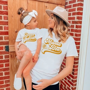 Pu&ograve; includere: Una maglietta bianca con il testo "Mama Bee" in un font giallo retr&ograve;. Un bambino indossa una maglietta bianca con il testo "Queen Bee" in un font giallo retr&ograve;. Entrambe le magliette presentano una piccola grafica di un'ape.