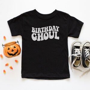 May include: Black t-shirt with the text "Birthday Ghoul" in white, a small orange pumpkin, and black and white sneakers on a white background.