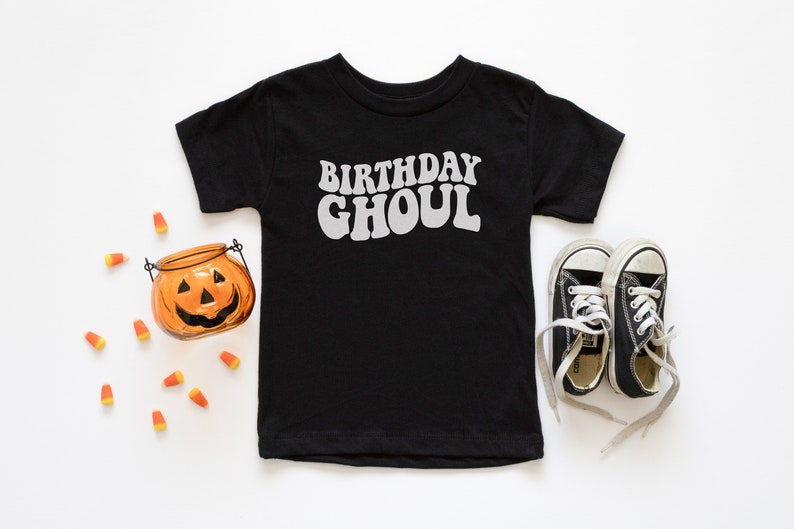 May include: Black t-shirt with the text "Birthday Ghoul" in white, a small orange pumpkin, and black and white sneakers on a white background.