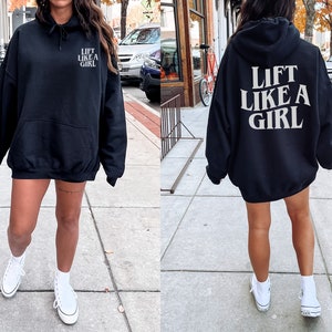 K&ouml;nnte beinhalten: Ein schwarzer Oversize-Hoodie mit dem Aufdruck "Lift Like A Girl" in Wei&szlig; auf der R&uuml;ckseite. Der Hoodie wird von einer Person mit wei&szlig;en Turnschuhen getragen.
