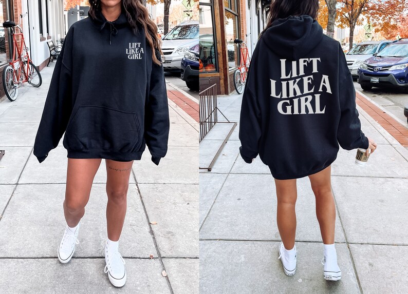 K&ouml;nnte beinhalten: Ein schwarzer Oversize-Hoodie mit dem Aufdruck "Lift Like A Girl" in Wei&szlig; auf der R&uuml;ckseite. Der Hoodie wird von einer Person mit wei&szlig;en Turnschuhen getragen.