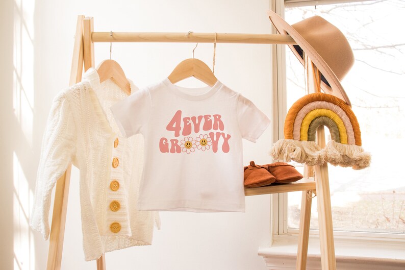 K&ouml;nnte beinhalten: Ein wei&szlig;es T-Shirt mit dem Text "4EVER GROOVY" in Rosa, mit G&auml;nsebl&uuml;mchen-Akzenten. Eine cremefarbene Strickjacke und ein brauner Hut sind ebenfalls zu sehen. Die Artikel werden auf einem Holzst&auml;nder mit einem Wandbehang in Regenbogenfarben pr&auml;sentiert.