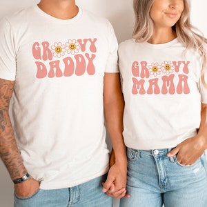 K&ouml;nnte beinhalten: Zwei cremefarbene T-Shirts mit den Worten "GROOVY DADDY" und "GROOVY MAMA" in Rosa, mit G&auml;nsebl&uuml;mchen-Akzenten. Die Shirts werden von zwei Personen getragen, die sich an den H&auml;nden halten. Das Set ist passend.