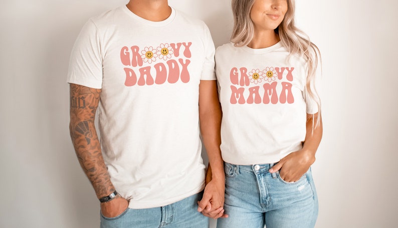K&ouml;nnte beinhalten: Zwei cremefarbene T-Shirts mit den Worten "GROOVY DADDY" und "GROOVY MAMA" in Rosa, mit G&auml;nsebl&uuml;mchen-Akzenten. Die Shirts werden von zwei Personen getragen, die sich an den H&auml;nden halten. Das Set ist passend.
