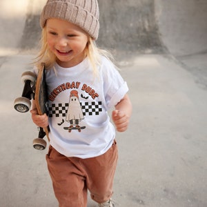 May include: A young person wearing a white t-shirt with a ghost graphic and the text "Birthday Dude" in orange. The person is holding a skateboard and wearing a brown beanie and brown pants.