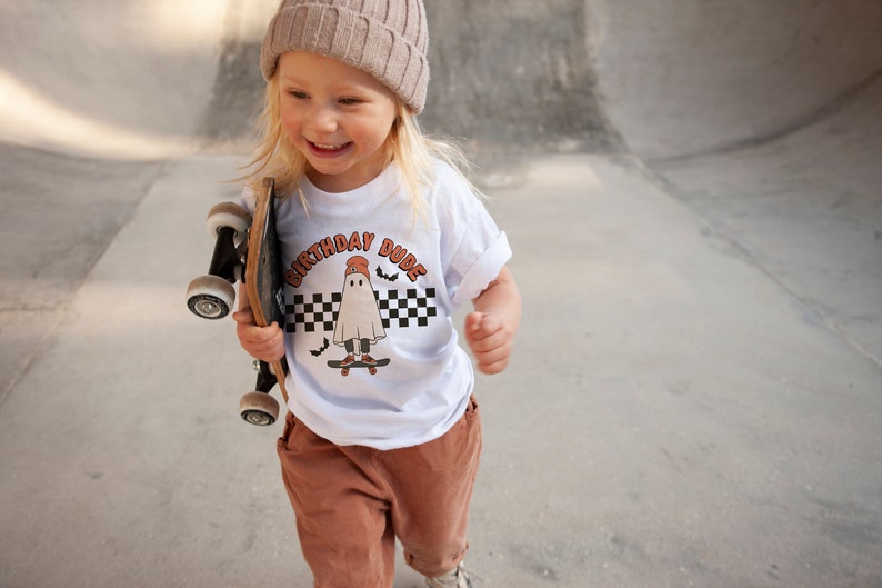May include: A young person wearing a white t-shirt with a ghost graphic and the text "Birthday Dude" in orange. The person is holding a skateboard and wearing a brown beanie and brown pants.