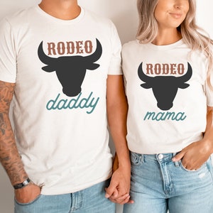 May include: A couple wearing white t-shirts with a black bull silhouette and the text "RODEO daddy" and "RODEO mama" in brown and blue.