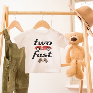 May include: White t-shirt with a red race car graphic and the text "two fast" in black. The shirt is hanging on a wooden rack with a pair of green overalls and a teddy bear.