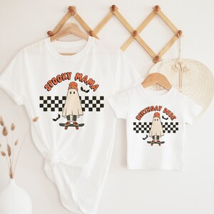 May include: Two white t-shirts with a ghost wearing a hat and skateboarding on a checkered background. The larger shirt says "Spooky Mama" and the smaller shirt says "Birthday Dude".