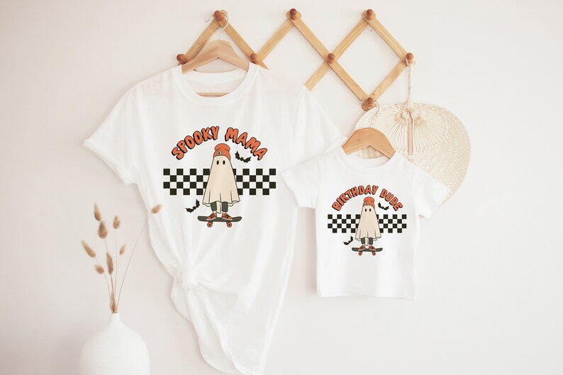 May include: Two white t-shirts with a ghost wearing a hat and skateboarding on a checkered background. The larger shirt says "Spooky Mama" and the smaller shirt says "Birthday Dude".