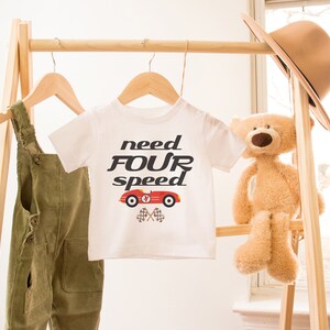 K&ouml;nnte beinhalten: Wei&szlig;es T-Shirt mit einem roten Rennwagen-Motiv und dem Text "need four speed". Das Shirt h&auml;ngt an einem Holzgestell mit einer gr&uuml;nen Latzhose und einem Teddyb&auml;r.