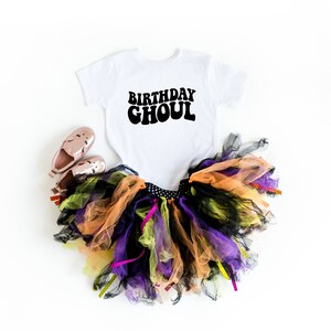 May include: A white onesie with the text "Birthday Ghoul" in black. The onesie is paired with a multi-colored tulle tutu skirt and pink shoes.