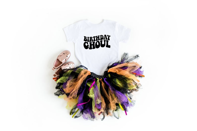 May include: A white onesie with the text "Birthday Ghoul" in black. The onesie is paired with a multi-colored tulle tutu skirt and pink shoes.