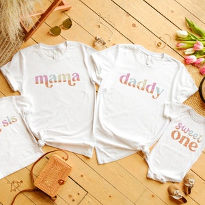 May include: Four white t-shirts with colorful text. The shirts say "mama", "daddy", "big sis", and "sweet one".
