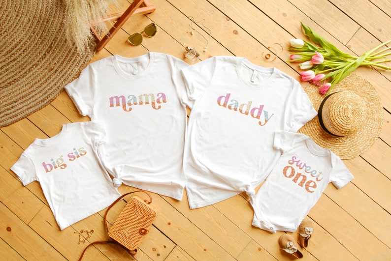 May include: Four white t-shirts with colorful text. The shirts say "mama", "daddy", "big sis", and "sweet one".