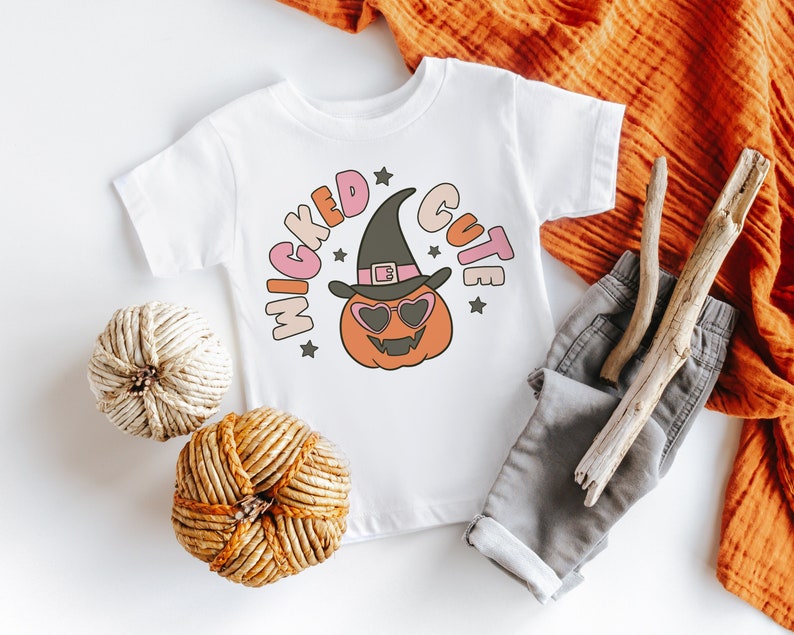 Puede incluir: Un body blanco para beb&eacute; con un gr&aacute;fico rosa, naranja y amarillo de una calabaza con un sombrero de bruja y gafas de sol. El texto "Wicked Cute" est&aacute; escrito alrededor de la calabaza.