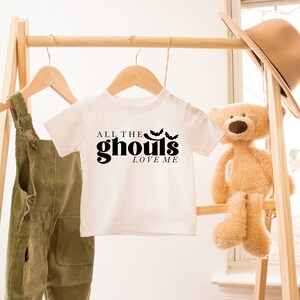 May include: White t-shirt with black text that reads "All the ghouls love me" and three black bat silhouettes. The shirt is hanging on a wooden rack with a pair of green overalls and a teddy bear.
