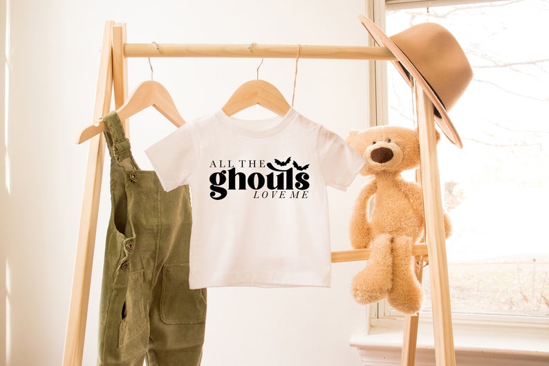 May include: White t-shirt with black text that reads "All the ghouls love me" and three black bat silhouettes. The shirt is hanging on a wooden rack with a pair of green overalls and a teddy bear.
