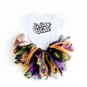 May include: A white bodysuit with the text "Spooky Babe" in black script with a spiderweb graphic. A black, orange, and purple tulle tutu with black and yellow ribbons. Two pink shoes.