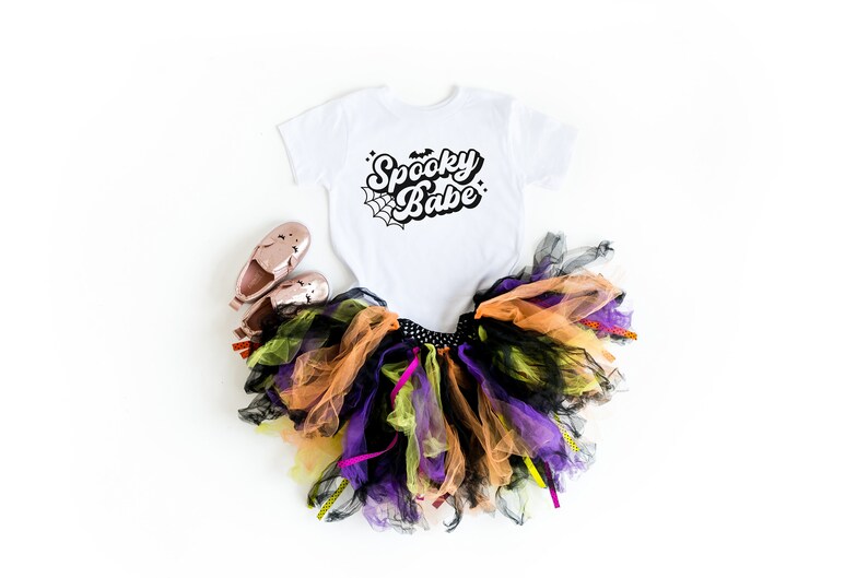May include: A white bodysuit with the text "Spooky Babe" in black script with a spiderweb graphic. A black, orange, and purple tulle tutu with black and yellow ribbons. Two pink shoes.