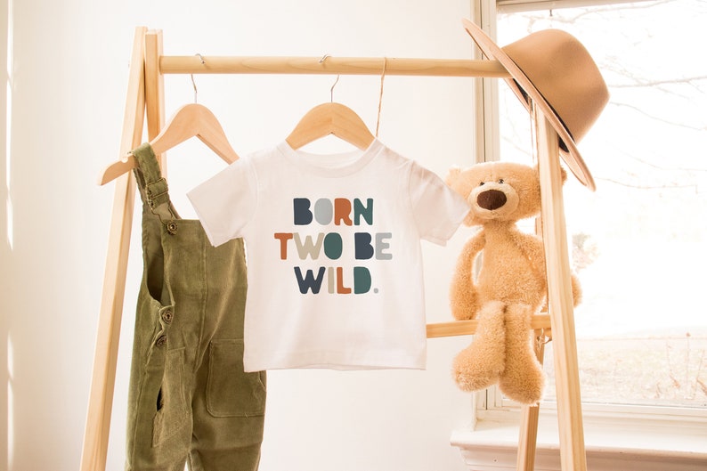 May include: White t-shirt with the text "BORN TWO BE WILD." in colorful block letters. The shirt is hanging on a wooden rack with a pair of green overalls and a stuffed teddy bear.