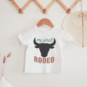 K&ouml;nnte beinhalten: Ein wei&szlig;er Baby-Strampler mit einer schwarzen Bullensilhouette und dem Text "my first rodeo" in T&uuml;rkis und Braun.