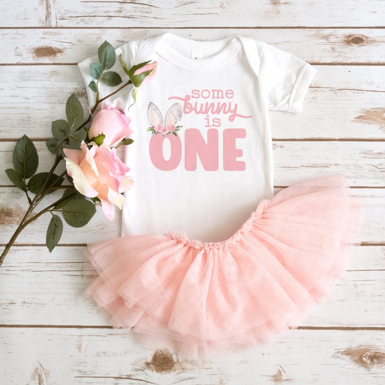 May include: A white onesie with pink bunny ears and flowers, and the text "some bunny is one". The onesie is paired with a pink tulle tutu.