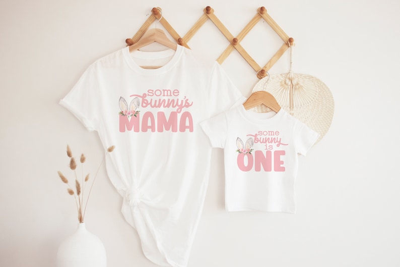 May include: Two white t-shirts with pink bunny graphics and text. The larger shirt says "some bunny's mama" and the smaller shirt says "some bunny is one".