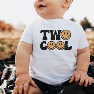 May include: A white toddler t-shirt with black text that reads "TWO COOL" with two yellow smiley faces. The toddler is wearing the shirt and sitting on a plaid blanket.