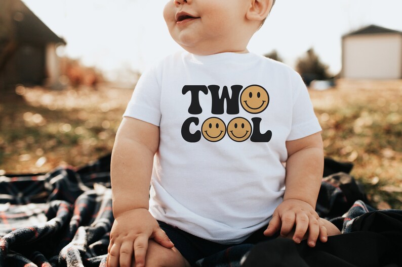 May include: A white toddler t-shirt with black text that reads "TWO COOL" with two yellow smiley faces. The toddler is wearing the shirt and sitting on a plaid blanket.