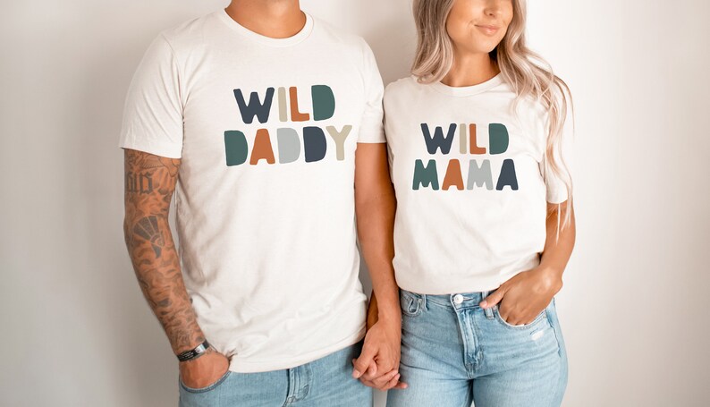 May include: Two white t-shirts with the text "WILD DADDY" and "WILD MAMA" in colorful block letters. The shirts are worn by two people holding hands.