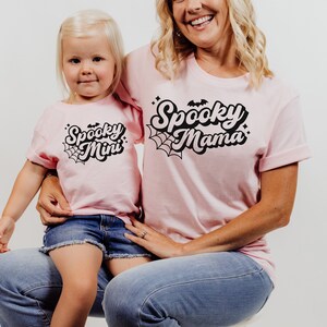 May include: A pink t-shirt with the text "Spooky Mama" in black with a spiderweb design. A smaller pink t-shirt with the text "Spooky Mini" in black with a spiderweb design.
