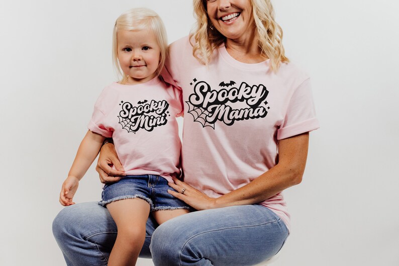 May include: A pink t-shirt with the text "Spooky Mama" in black with a spiderweb design. A smaller pink t-shirt with the text "Spooky Mini" in black with a spiderweb design.