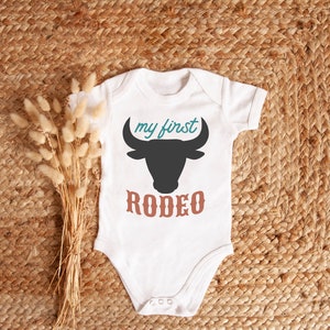 May include: White baby onesie with a black bull silhouette and the text "my first rodeo" in teal and brown.
