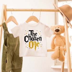 May include: White baby onesie with black and gold text that reads "The Chosen One" with a gold lightning bolt graphic. The onesie is hanging on a wooden rack with a teddy bear and a hat.