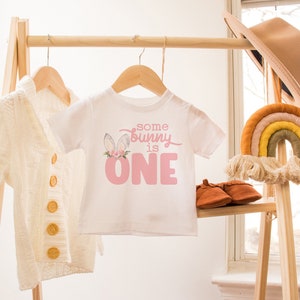 May include: A white baby onesie with a pink bunny graphic and the text "some bunny is one". The onesie is hanging on a wooden rack with a white cardigan and a rainbow macrame hanging on the right.