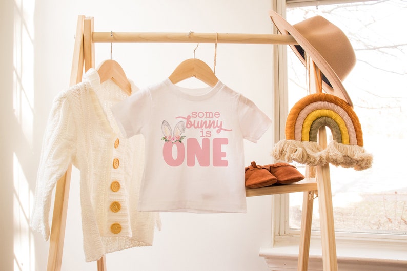 May include: A white baby onesie with a pink bunny graphic and the text "some bunny is one". The onesie is hanging on a wooden rack with a white cardigan and a rainbow macrame hanging on the right.