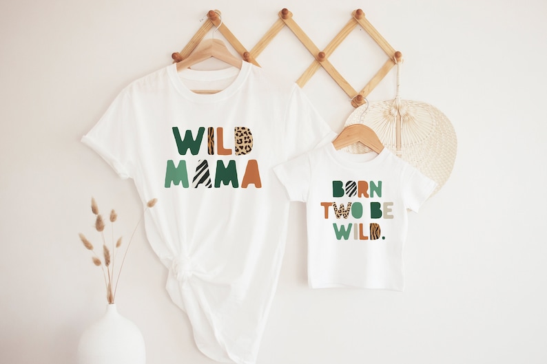 May include: Two white t-shirts with a green, orange, and brown animal print design. The larger shirt says "WILD MAMA" and the smaller shirt says "BORN TO BE WILD."