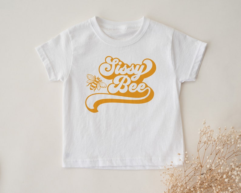Pu&ograve; includere: Maglietta bianca a maniche corte con una grafica gialla in stile retr&ograve; che dice "Sissy Bee" e una piccola illustrazione di un'ape.