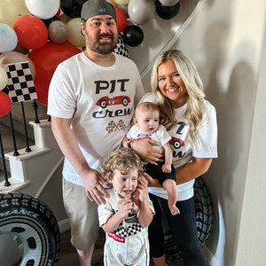 May include: A family of four poses for a photo. The parents are wearing white t-shirts with a red race car graphic and the text "PJT Crew". The older child is wearing a white racing suit with a checkered pattern and the text "Goodyear". The baby is wearing a white onesie with a red race car graphic. The family is standing in front of a wall decorated with balloons and checkered flags.