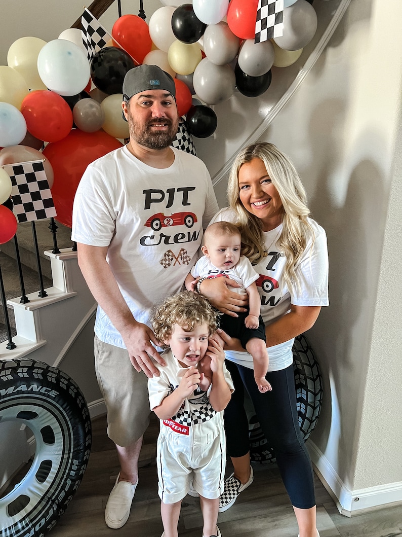 May include: A family of four poses for a photo. The parents are wearing white t-shirts with a red race car graphic and the text "PJT Crew". The older child is wearing a white racing suit with a checkered pattern and the text "Goodyear". The baby is wearing a white onesie with a red race car graphic. The family is standing in front of a wall decorated with balloons and checkered flags.
